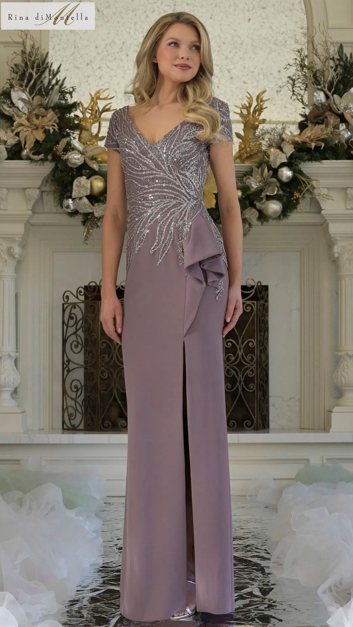 A woman poses indoors in the Rina Di Montella RD3323 Long Formal Dress in Victorian Lilac, featuring a beaded bodice, short sleeves, front slit, and decorative ruffle, set against a festively decorated background.